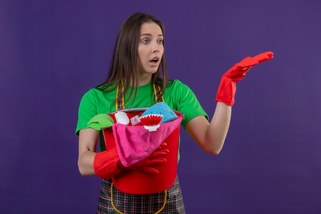 Regardant à côté surpris nettoyage jeune femme en uniforme dans des gants rouges tenant des outils de nettoyage levant la main sur un mur violet isolé