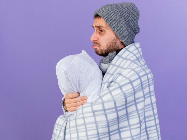 Regardant à côté triste jeune homme malade portant un chapeau d'hiver avec un foulard enveloppé dans un oreiller à carreaux et étreint isolé sur fond violet