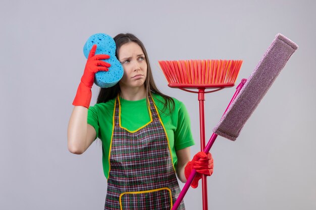 Regardant le côté triste nettoyage jeune femme en uniforme dans des gants rouges tenant des outils de nettoyage sur mur blanc isolé