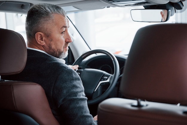 En regardant de côté. Vue de derrière d'homme d'affaires senior en tenue officielle conduisant une nouvelle voiture moderne