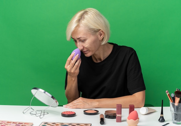 En regardant le miroir, une belle jeune fille est assise à table avec des outils de maquillage appliquant une crème tonifiante avec une éponge isolée sur un mur vert