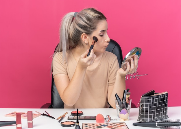 En regardant le miroir, une belle jeune fille est assise à table avec des outils de maquillage appliquant un fard à joues en poudre isolé sur un mur rose