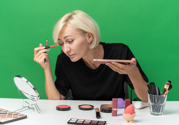 En regardant le miroir, une belle jeune fille est assise à table avec des outils de maquillage appliquant un fard à paupières avec un pinceau de maquillage isolé sur un mur vert