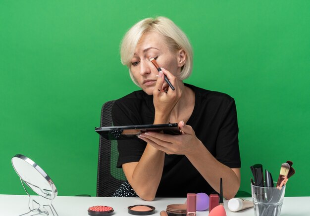 En regardant le miroir, une belle jeune fille est assise à table avec des outils de maquillage appliquant un fard à paupières avec un pinceau de maquillage isolé sur un mur vert