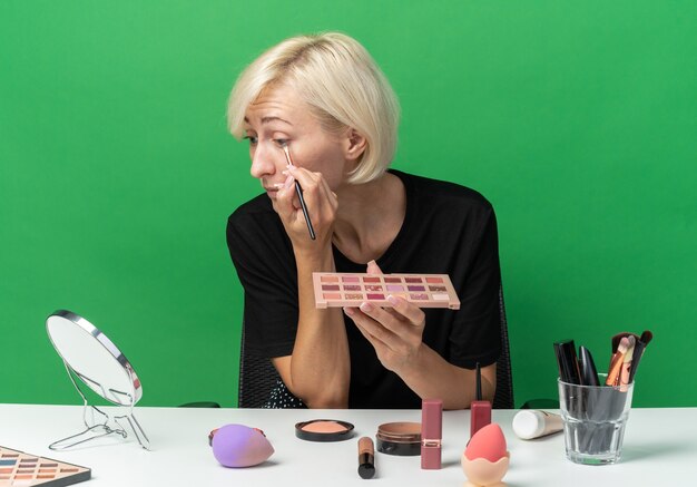 En regardant le miroir, une belle jeune fille est assise à table avec des outils de maquillage appliquant un fard à paupières avec un pinceau de maquillage isolé sur un mur vert