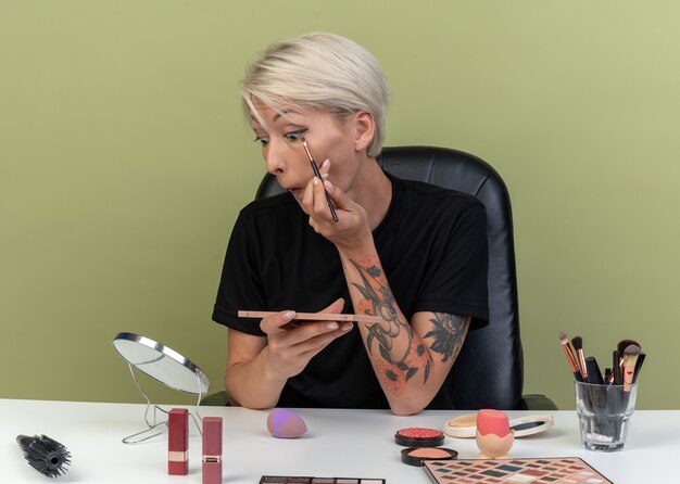 En regardant le miroir, une belle jeune fille est assise à table avec des outils de maquillage appliquant un fard à paupières avec des pinceaux de maquillage isolés sur un mur vert olive