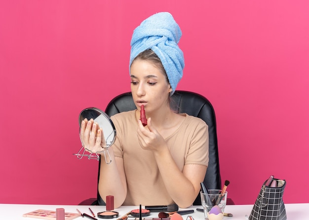 En regardant le miroir, une belle jeune fille est assise à table avec des outils de maquillage enveloppés dans une serviette en appliquant du rouge à lèvres isolé sur un mur rose