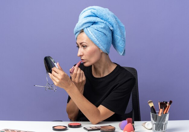 En regardant le miroir, une belle jeune fille est assise à table avec des outils de maquillage en essuyant les cheveux dans une serviette en appliquant du rouge à lèvres isolé sur un mur bleu