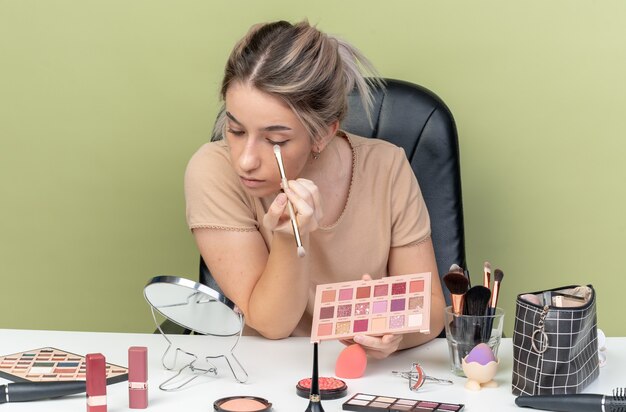 Regardant le miroir jeune belle fille assise au bureau avec des outils de maquillage appliquant un fard à paupières avec un pinceau de maquillage isolé sur un mur vert olive