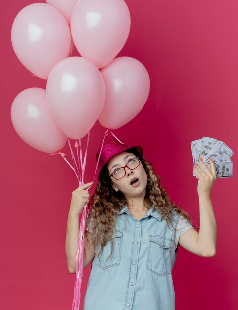 Regardant la pensée de la jeune fille portant des lunettes et un chapeau rose tenant des ballons et de l'argent isolé sur fond rose