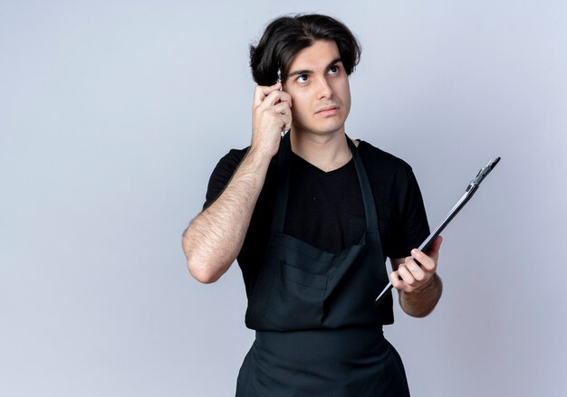 Regardant la pensée de jeune homme beau coiffeur en uniforme tenant le presse-papiers et mettant un stylo sur le front isolé sur fond blanc