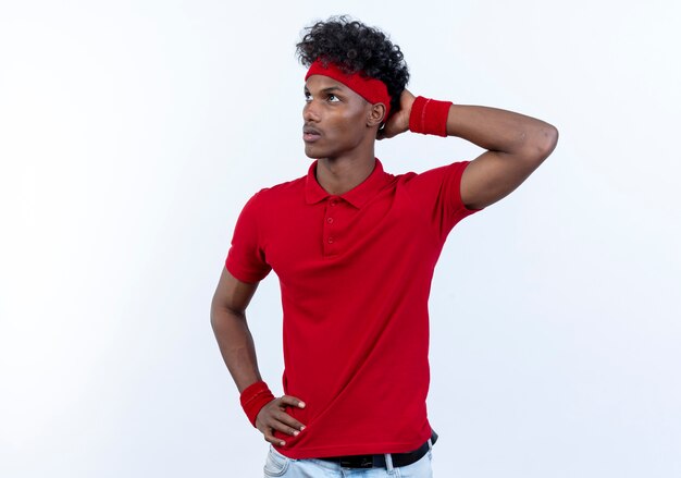 Regardant la pensée de jeune homme sportif afro-américain portant bandeau et bracelet mettant la main sur la tête isolé sur fond blanc