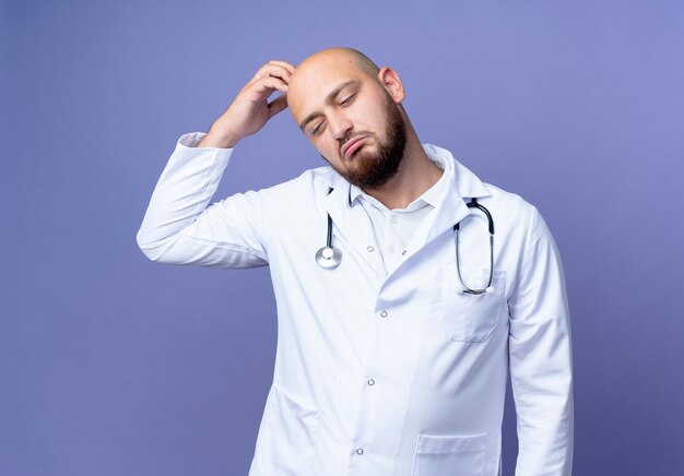 Regardant vers le bas confus jeune médecin de sexe masculin chauve portant robe médicale et stéthoscope se gratter la tête isolé sur fond bleu