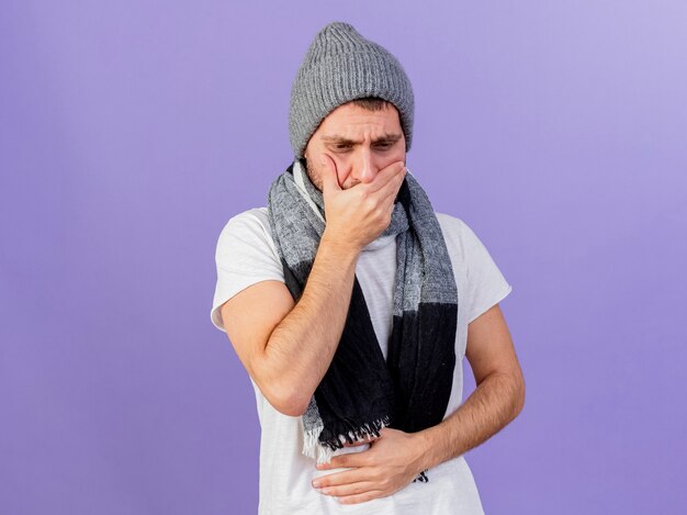 Regardant vers le bas jeune homme malade portant un chapeau d'hiver avec écharpe tenant la main sur mout et attrapé le ventre douloureux isolé sur fond violet