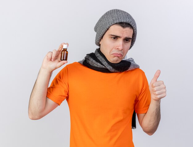 Regardant vers le bas triste jeune homme malade portant un chapeau d'hiver avec écharpe tenant des médicaments dans une bouteille en verre montrant le pouce vers le haut isolé sur fond blanc