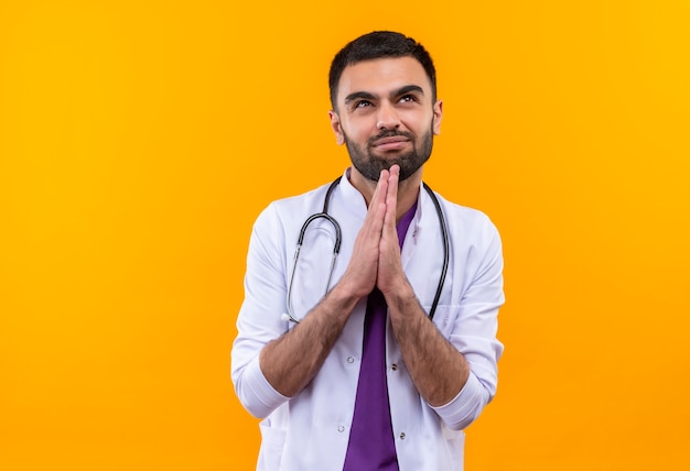Regardant vers le haut concerné jeune médecin de sexe masculin portant une robe médicale stéthoscope montrant le geste de prier sur fond jaune isolé