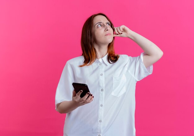 Regardant vers le haut confus jeune fille rousse tenant le téléphone et mettant le doigt sur la tête isolé sur fond rose