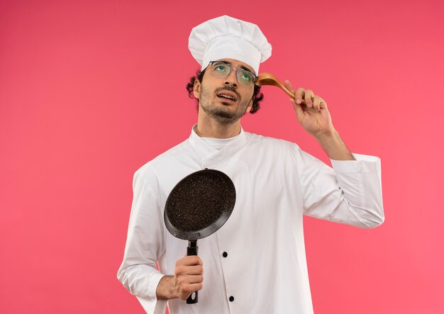 Regardant vers le haut confus jeune homme cuisinier portant l'uniforme de chef et des lunettes tenant une poêle et mettant une cuillère sur le front