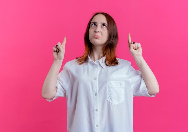 Regardant vers le haut impressionné jeune fille rousse pointe vers le haut isolé sur un mur rose