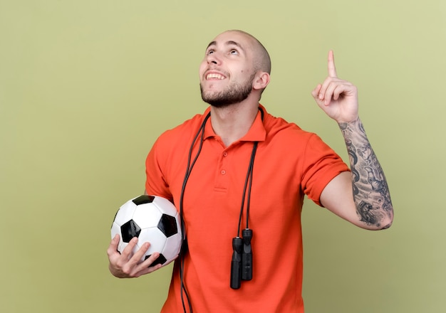Regardant vers le haut souriant jeune homme sportif pointe vers le haut tenant la balle avec corde à sauter sur l'épaule isolé sur fond vert olive