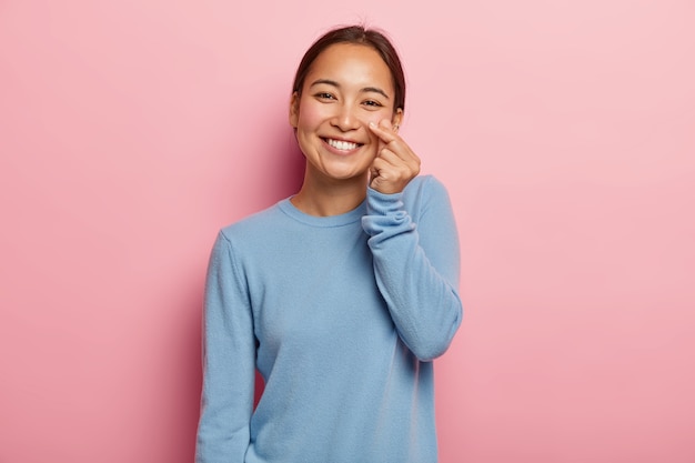 Regarde ma joue. Agréable femme asiatique touche la peau du visage, démontre sa fraîcheur et sa douceur, ne porte pas de maquillage, cheveux peignés foncés, vêtue d'un sweat-shirt bleu décontracté, isolé sur rose