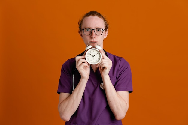 Regarder La Caméra Tenant Le Réveil Jeune Homme Médecin Portant L'uniforme Avec Stéthoscope Isolé Sur Fond Orange