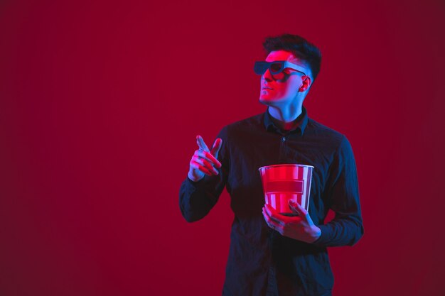 Regarder à travers des lunettes 3D. Portrait d'un jeune homme caucasien isolé sur un mur rouge à la lumière du néon. Beau modèle. Concept d'émotions humaines, d'expression faciale, de jeunesse, d'appareils.