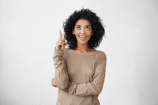 Regardez ça! Portrait intérieur de joyeuse jolie jeune femme habillée avec désinvolture aux cheveux bouclés pointant son index vers le haut, indiquant quelque chose d'intéressant, ayant un regard heureux et excité