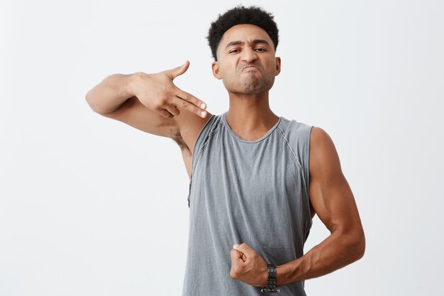 Regardez ce corps. Portrait de jeune homme sérieux à la peau sombre avec une coiffure afro montrant le geste du pistolet avec la main, pointant ses muscles avec une expression moyenne du visage.