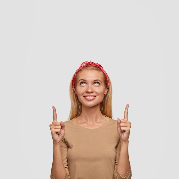 Regardez cette direction. Belle jeune femme blonde joyeuse avec un large sourire doux, pointe vers le haut, étant à quelque chose de drôle, se tient contre un mur blanc, vêtue de vêtements décontractés.