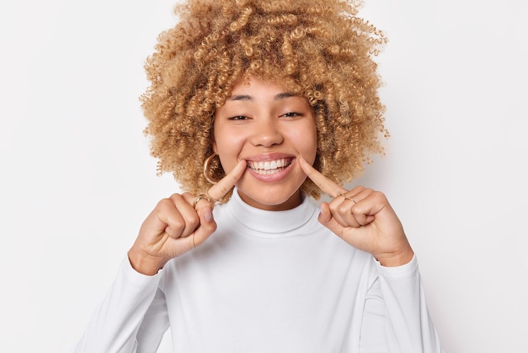 Photo gratuit regardez mon sourire orthodontique parfait. une femme positive avec des cheveux bouclés et touffus des points d'expression gais sur des dents blanches sourit largement prend soin de la santé dentaire pose à l'intérieur exprime des émotions heureuses