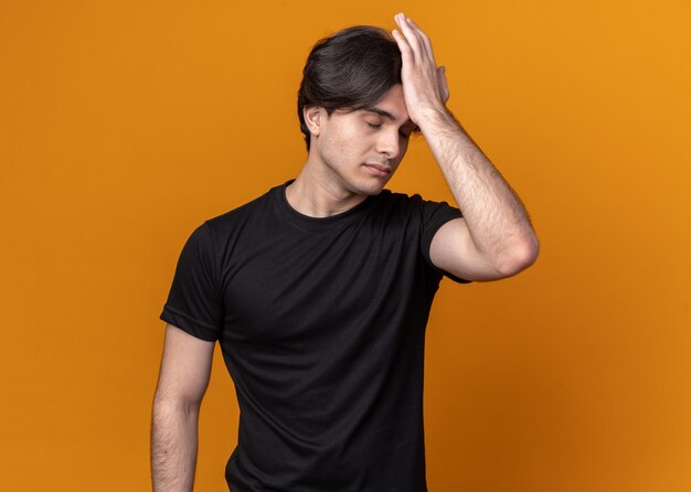 Regretté jeune beau mec portant un t-shirt noir mettant la main sur le front isolé sur un mur orange