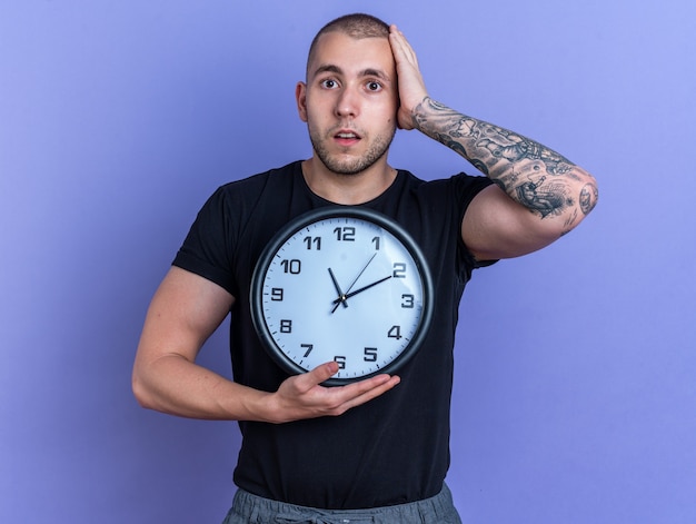 Regretté jeune beau mec portant un t-shirt noir tenant une horloge murale mettant la main sur la tête isolée sur le mur bleu