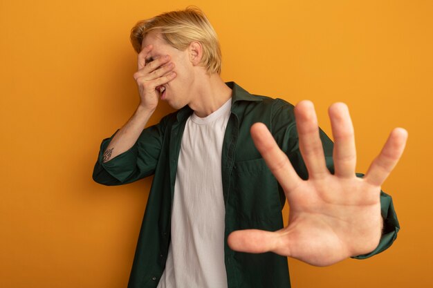 Regretté jeune mec blond portant un t-shirt vert visage couvert avec la main et tenant la main