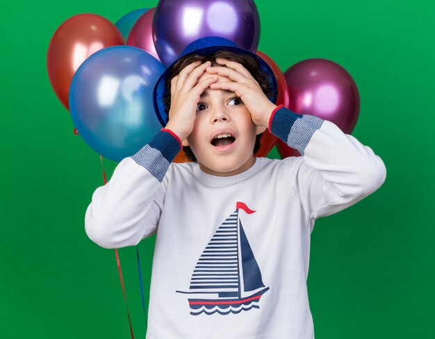Regretté petit garçon portant un chapeau de fête bleu debout devant des ballons attrapé la tête