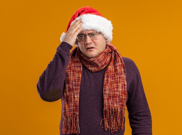 Regretting homme adulte portant des lunettes et bonnet de Noel avec foulard autour du cou mettant la main sur la tête à côté isolé sur fond orange