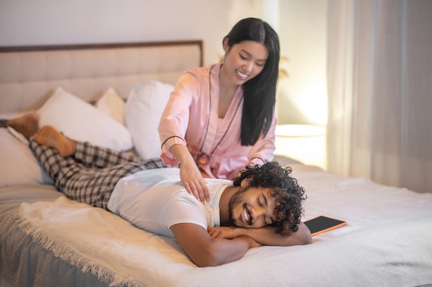 Relaxant. Jeune adulte femme asiatique aux cheveux longs en pyjama rose assis donnant un massage du dos à un homme indien souriant allongé sur le lit dans la chambre