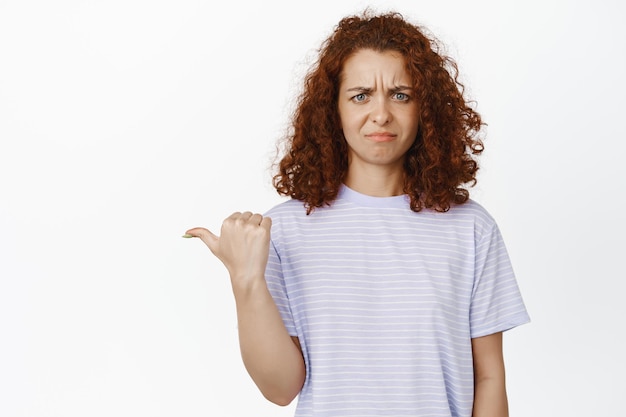 Photo gratuite répugnant. une jeune femme rousse mécontente grimace, pointe à gauche avec aversion et aversion, se tient en t-shirt sur fond blanc