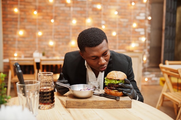 Respectable jeune homme afro-américain en costume noir et gants pour la nourriture assis au restaurant avec un délicieux double burger et une boisson gazeuse