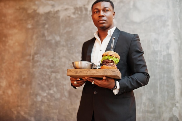 Respectable jeune homme afro-américain en costume noir tenir le plateau avec double burger contre le mur gris