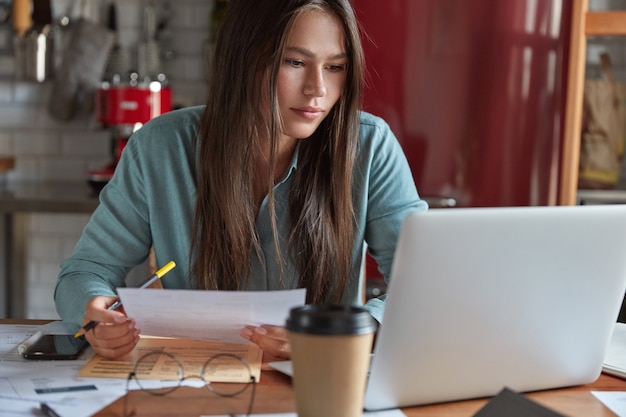 Un responsable commercial sérieux a concentré son expression sur l'écran d'un ordinateur portable, fait un rapport comptable sur les ventes, fait un travail à distance à la maison, pose à la cuisine, entouré de documents papier et de boissons savoureuses