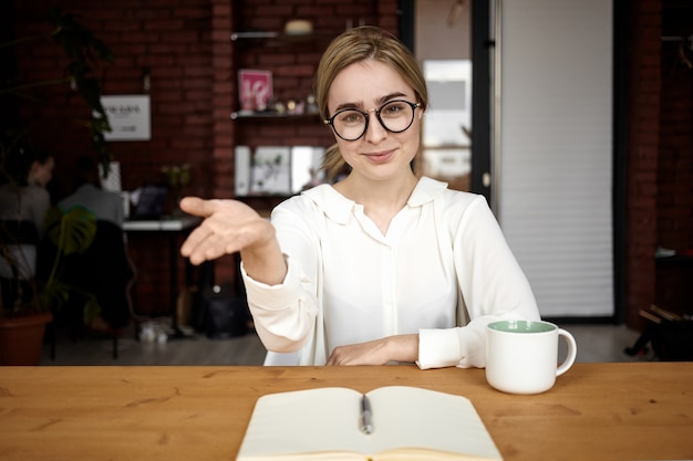 Responsable des ressources humaines polie en lunettes, assise à son bureau, tendant la main à la caméra, ouverte à la coopération, faisant signe de bienvenue, disant: Veuillez prendre place. Partenaire d'accueil de femme d'affaires sympathique