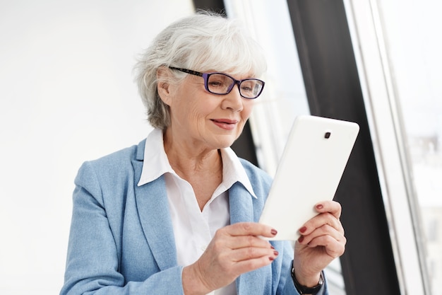 Rester connecté. Femme âgée intelligente moderne avec des cheveux gris posant isolé dans des lunettes et des vêtements formels, lisant un livre électronique ou faire du shopping en ligne à l'aide d'une tablette numérique, ayant heureux regard heureux