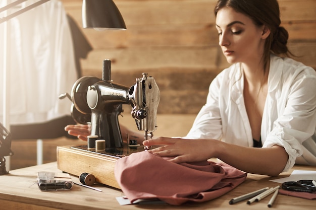 Photo gratuite restez calme et cousez avec passion. plan intérieur d'une femme travaillant avec du tissu sur une machine à coudre, essayant de se concentrer en atelier. jeune créatrice faisant un nouveau vêtement pour son amie