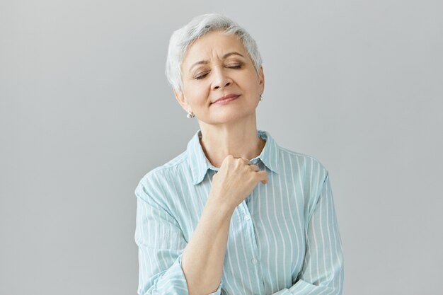 Retraité belle femme aux cheveux gris en chemise rayée bleue fermant les yeux et souriant paisiblement, profitant de la bonne musique classique, ayant une expression faciale nostalgique, tenant sa poitrine