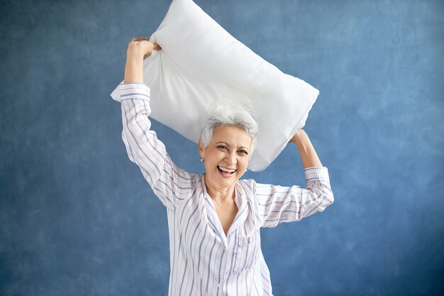 Retraité femme portant un pyjama soyeux rire, être de bonne humeur tout en s'amusant dans la chambre, en levant les bras, tenant un oreiller en plumes au-dessus de sa tête