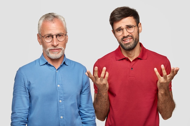 Un retraité masculin attrayant coopère avec son jeune collègue qui a une expression nerveuse désespérée, debout l'un à côté de l'autre, isolé sur un mur blanc. Les gens et les relations