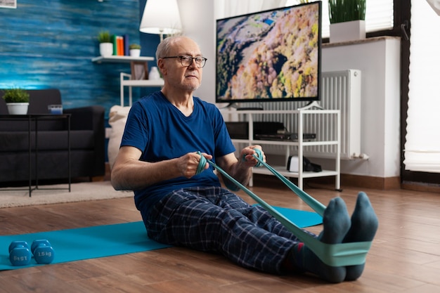 Retraite senior homme assis sur un tapis de yoga étirant les muscles des jambes à l'aide d'une bande élastique de résistance formant la flexibilité du corps. Retraité en sportswear amincissant le poids pendant l'entraînement musculaire dans le salon