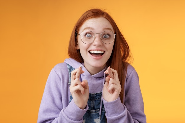 Un rêve de fille surprise est devenu réalité célébrant debout amusé excité croiser les doigts bonne chance souhait réalisé souriant émotif ravi enfin gagner recevoir de bonnes nouvelles optimiste optimiste fond orange