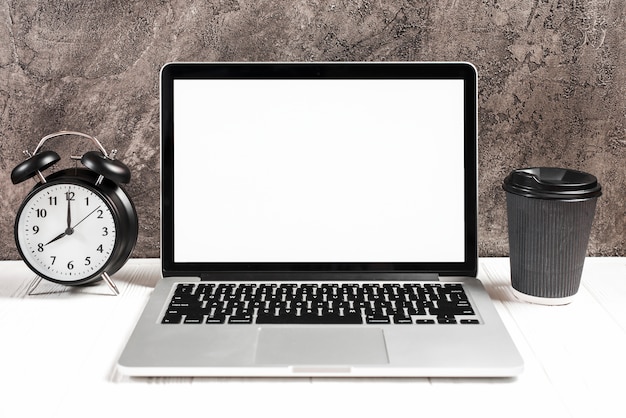 Réveil et tasse à café jetable avec un ordinateur portable ouvert sur un bureau blanc